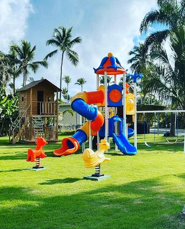 Bright tropical playground with colorful blue and orange tube slides, a wooden playhouse and spring ride-on toys on a green lawn under palm trees.