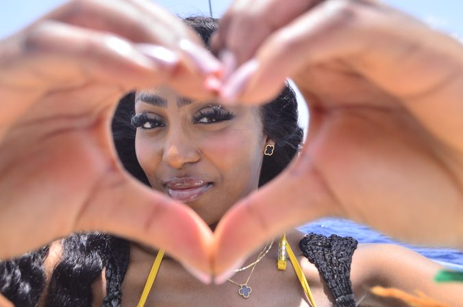 Close-up beach portrait of a smiling woman framing her face with heart-shaped hands, ocean and sunny sky in the background, wearing a yellow bikini strap, black cover-up and a clover pendant