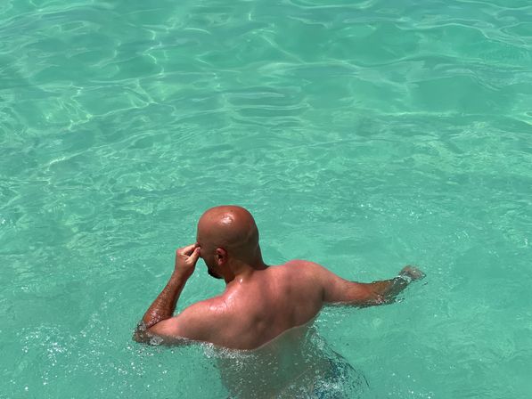 Bald swimmer pinching his nose while wading in clear turquoise water, sunlit ripples and gentle waves