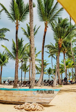 Wooden boat and coiled rope on a sandy tropical beach framed by tall palm trees, rows of lounge chairs and visitors with turquoise ocean waves in the background.