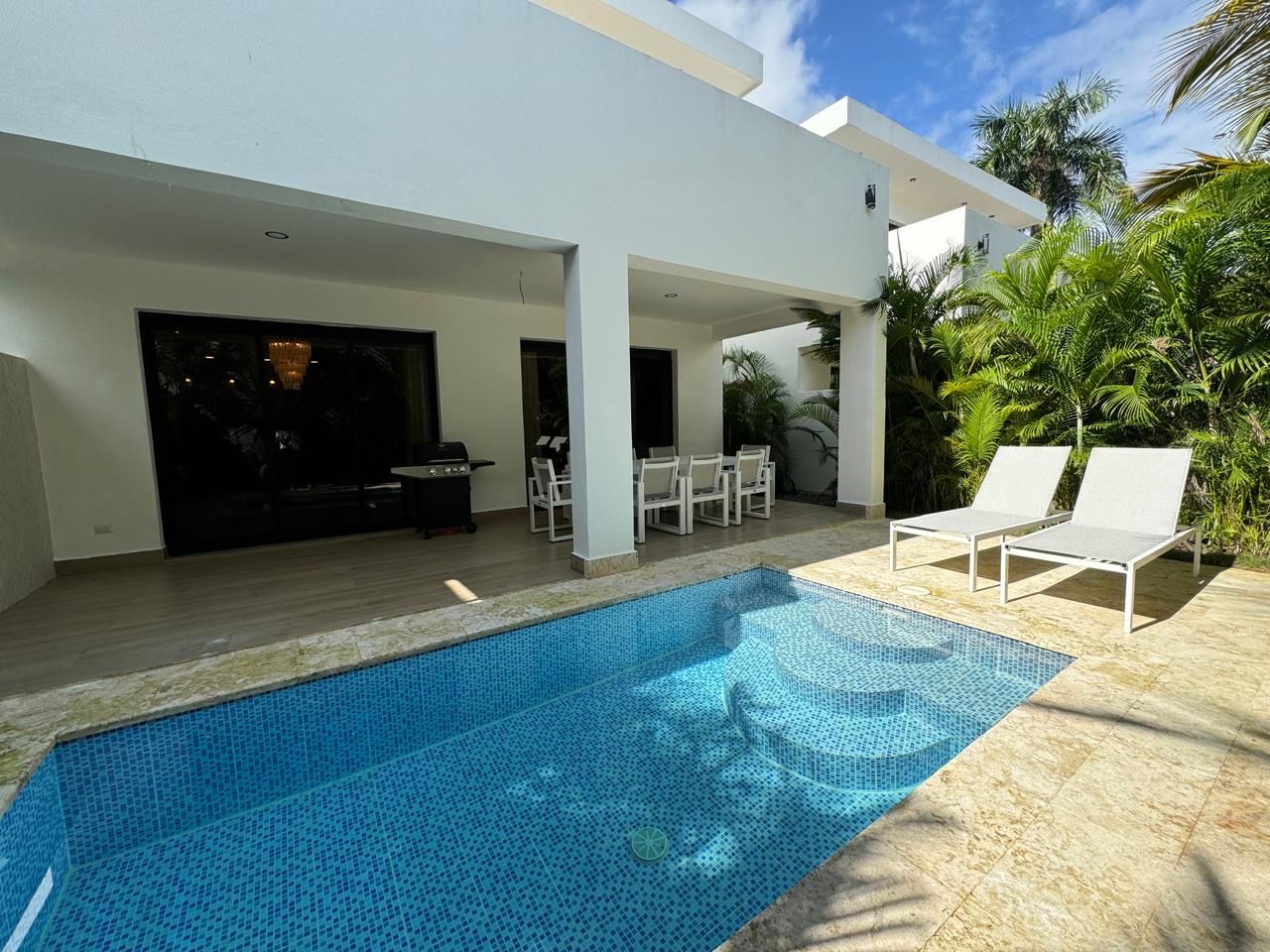 Modern white villa patio with blue-tiled plunge pool, stone deck, two lounge chairs, covered outdoor dining area and grill framed by tropical palm greenery under a sunny sky.