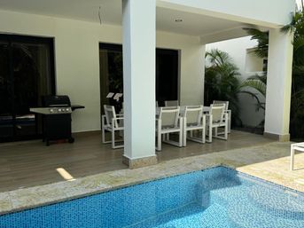 Modern covered patio at a tropical villa with a white outdoor dining set, gas grill, sliding glass doors and a blue mosaic-tiled swimming pool framed by palm plants