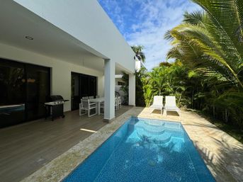 Sunlit tropical villa patio with blue-tiled plunge pool, covered white outdoor dining set and grill, two white sun loungers on a stone deck surrounded by palm trees and lush greenery.