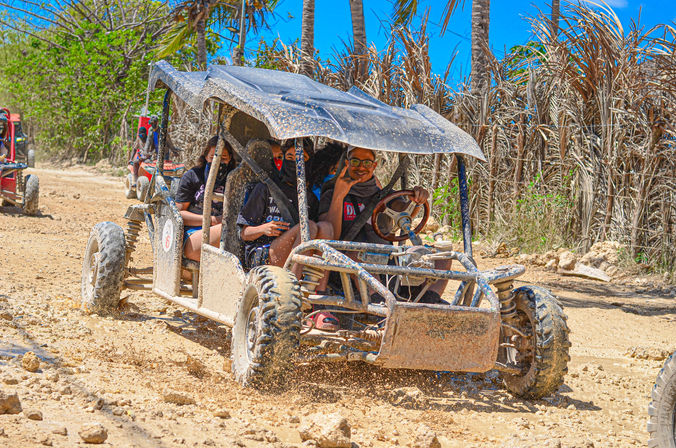 Smiling group enjoying a muddy off-road dune buggy ride along a tropical island dirt trail with palm trees and bright blue sky