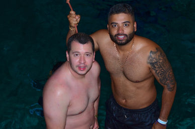 Two shirtless men smiling during a night swim in a pool, one holding a rope while the other displays a detailed arm tattoo, gold chain and wristband — friends enjoying a pool outing.
