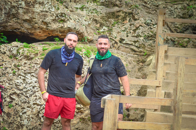 Two hikers in black shirts and colorful bandanas (blue, green) stand by a limestone rock formation and wooden stairs near a cave entrance, one carrying a duffel bag.
