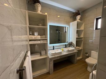 Bright modern marble bathroom with double vessel sinks, LED backlit mirror, open shelving of white towels, walk-in shower and wood-look floor.