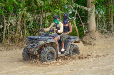 Two people on a black ATV splashing through muddy water on an off-road tropical forest trail, wearing bandanas and caps for a playful quad-bike adventure.