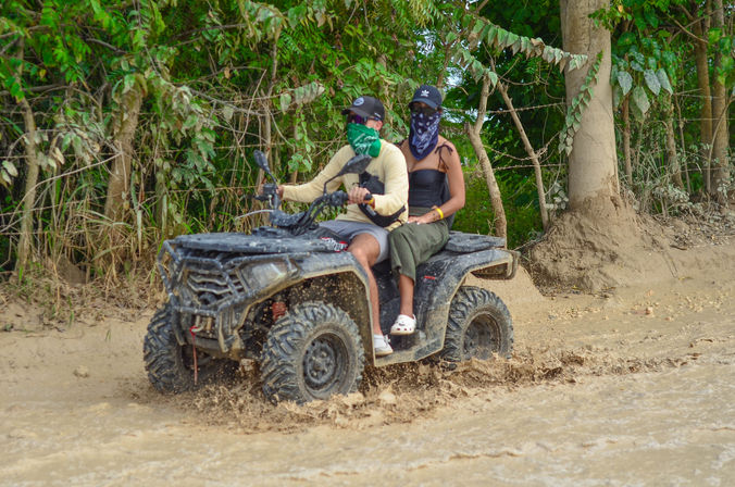 Two people on a black ATV splashing through muddy water on an off-road tropical forest trail, wearing bandanas and caps for a playful quad-bike adventure.