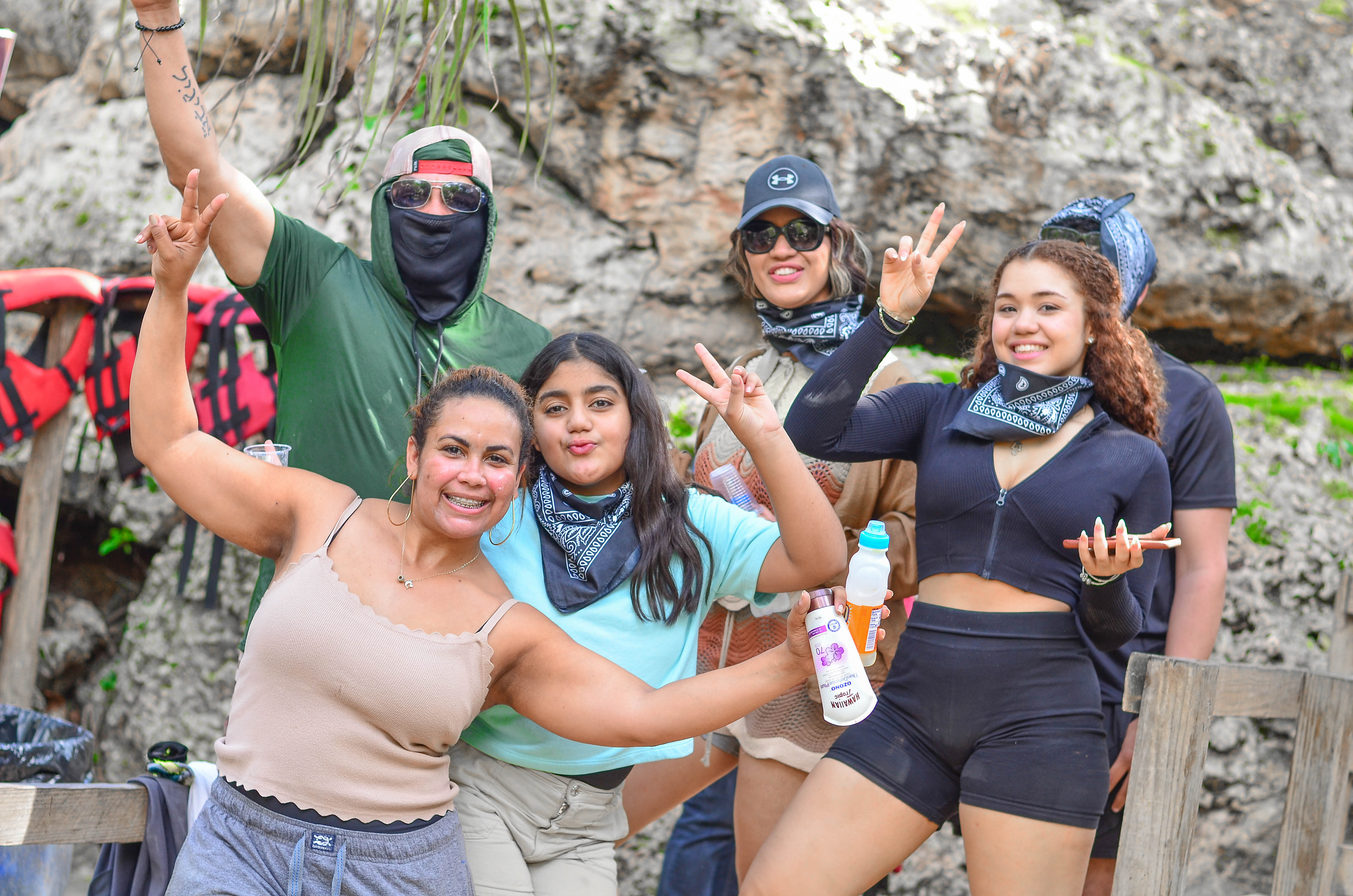 Smiling group of friends in bandanas and activewear posing with peace signs at a rocky riverside adventure spot, life jackets visible nearby