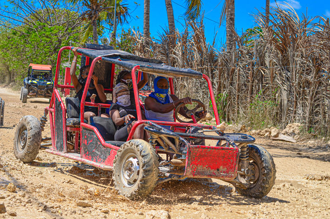 Red off-road dune buggy with four masked riders splashing mud on a sunny tropical dirt trail lined with palm trees