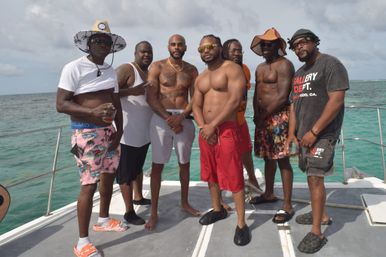 Seven men posing on a sunlit boat over turquoise tropical ocean, wearing swim trunks, hats and sunglasses and holding drinks — relaxed beach vacation vibe.