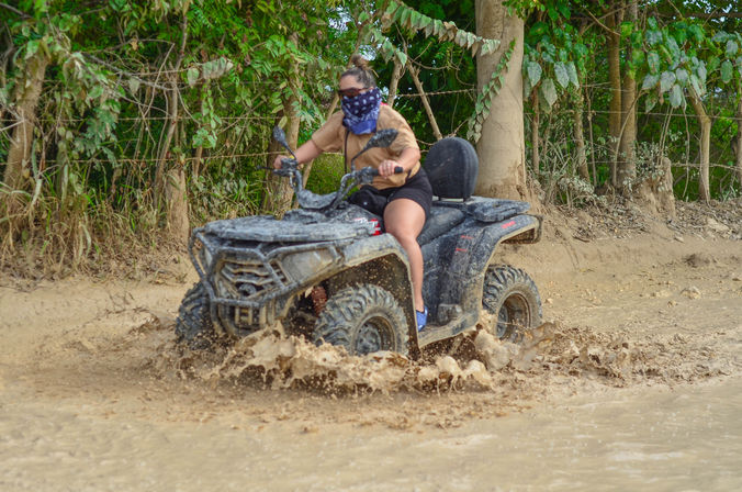 Rider wearing a bandana and sunglasses on an ATV splashing through a deep muddy puddle on a tropical dirt trail lined with trees.