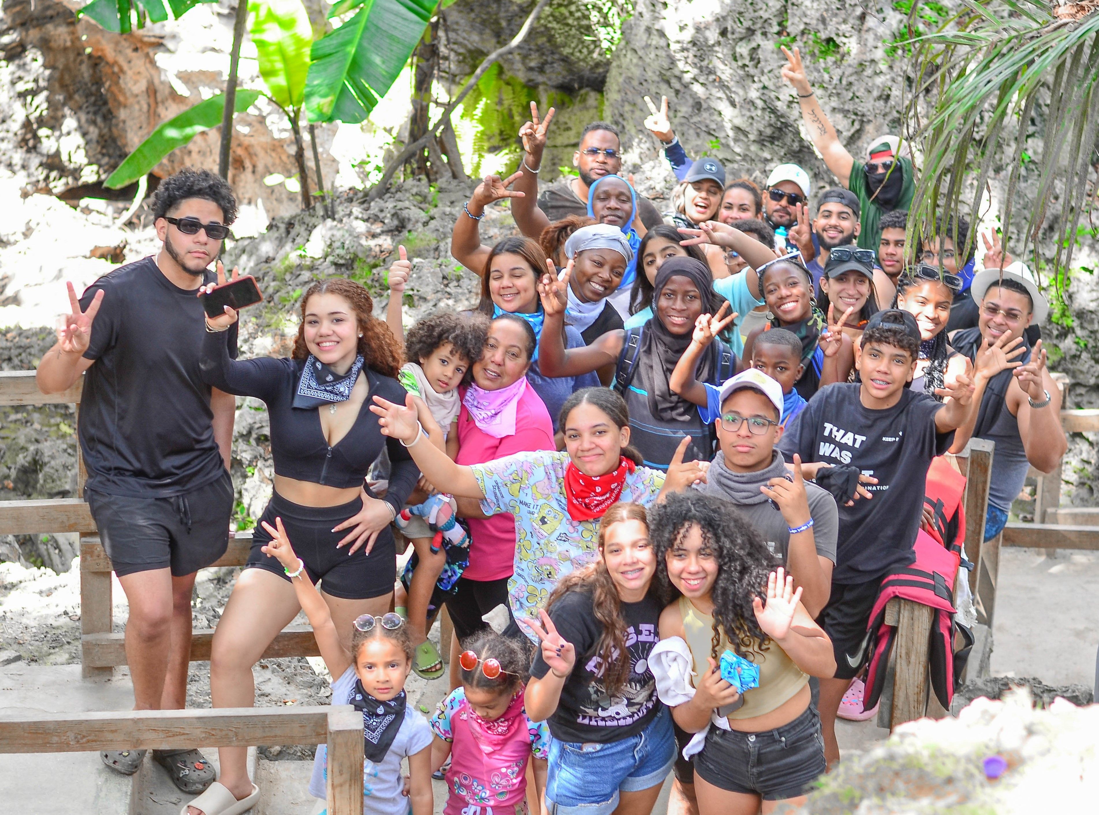 Smiling multigenerational group photo on a wooden trail in a tropical rock-and-jungle setting, friends and family posing with peace signs during an outdoor adventure/tropical hike.