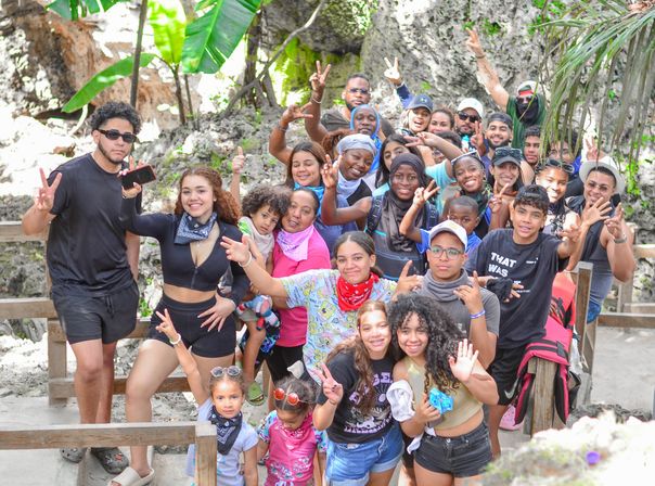Smiling multigenerational group photo on a wooden trail in a tropical rock-and-jungle setting, friends and family posing with peace signs during an outdoor adventure/tropical hike.