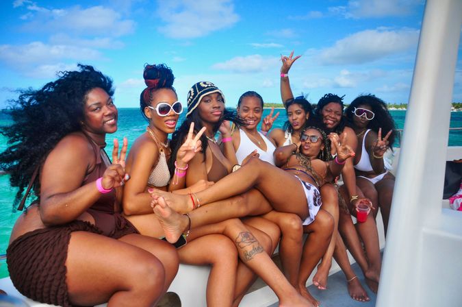 Eight women in swimsuits smiling and flashing peace signs on a boat over turquoise tropical ocean under a sunny sky
