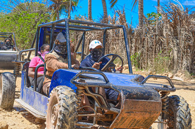 Four people wearing caps and bandanas riding a blue off-road dune buggy on a sandy tropical trail with palm trees and a bright blue sky.