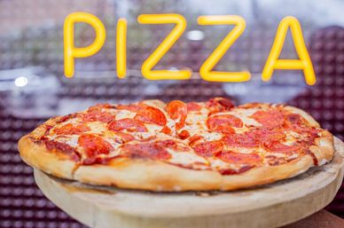 Mouthwatering pepperoni pizza on a wooden stand displayed in a pizzeria window under a glowing yellow neon 'PIZZA' sign