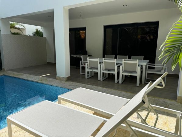 Sunlit tropical pool patio with blue mosaic pool, two white lounge chairs at the water’s edge and a modern outdoor dining set under a covered terrace.