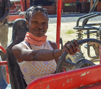 Smiling woman behind the wheel of a red off-road dune buggy on a sunny sandy track, hands on the steering wheel ready for outdoor adventure.