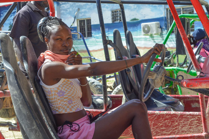 Woman seated in a red open-frame dune buggy with hand on the steering wheel, wearing a checkered crop top and pink shorts, parked in a sandy, beach-side lot with a mural of the ocean in the background — coastal off-road adventure.