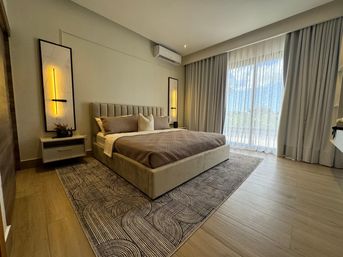 Modern master bedroom interior with beige upholstered king bed, patterned area rug, vertical wall sconces, air conditioner, and floor-to-ceiling sheer curtains over sliding doors letting in natural light.