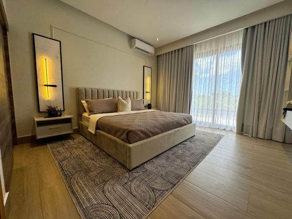 Modern master bedroom interior with beige upholstered king bed, patterned area rug, vertical wall sconces, air conditioner, and floor-to-ceiling sheer curtains over sliding doors letting in natural light.