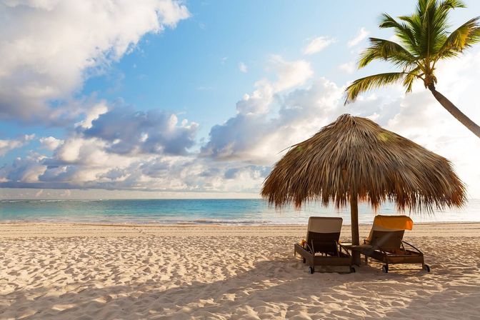 Sunlit tropical white-sand beach with calm turquoise water, two empty lounge chairs under a palm-frond thatched umbrella and a leaning palm tree beneath a partly cloudy sky.
