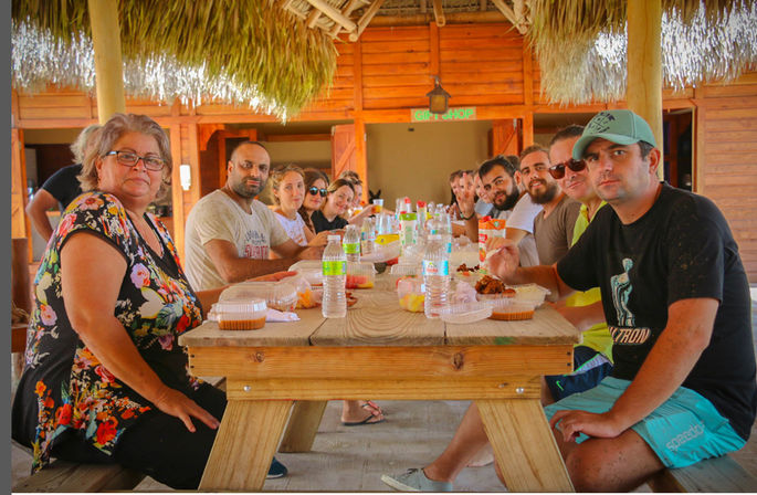 Smiling group enjoying a casual outdoor lunch at a long wooden picnic table under a thatched-roof palapa, with bottled water and takeout containers in a tropical seaside setting