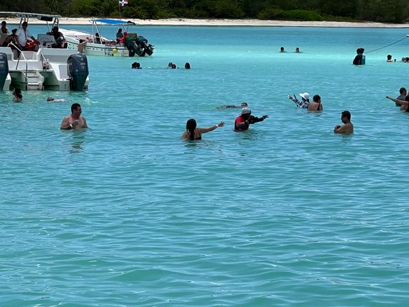 Vacationers wading and swimming in shallow turquoise tropical water near anchored motorboats and a sandy, tree-lined shore.