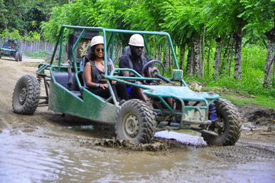 Two people in safety helmets splash through a muddy puddle in a green off-road dune buggy on a forest track surrounded by lush tropical trees, outdoor adventure scene.