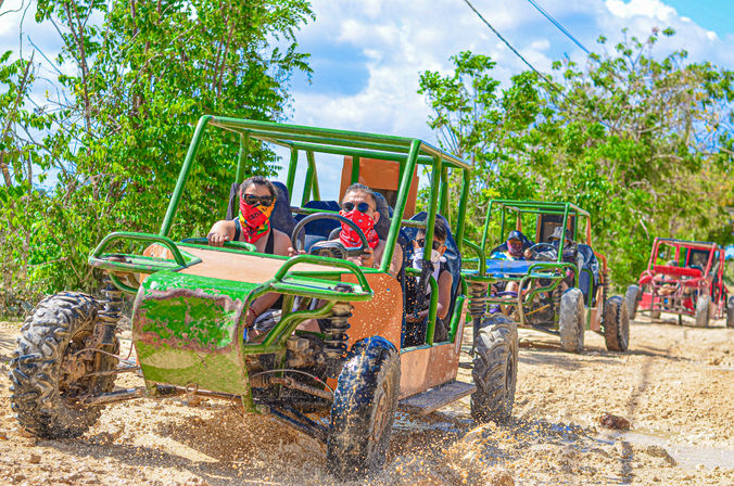 Off-road dune buggy tour on a muddy tropical trail, colorful buggies splashing through puddles with passengers wearing bandanas and sunglasses, green foliage and blue sky in the background