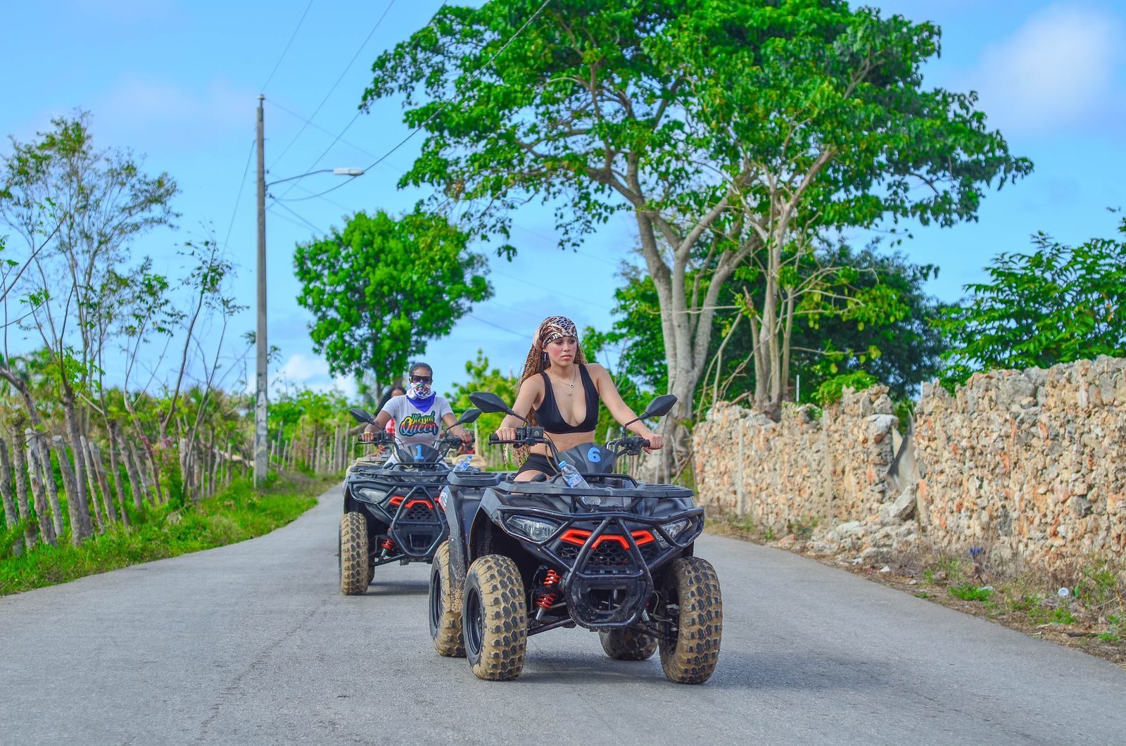 Two riders on black ATVs cruising a sunny tropical country road lined with a stone wall and lush green trees; lead rider wearing a bandana and black top.