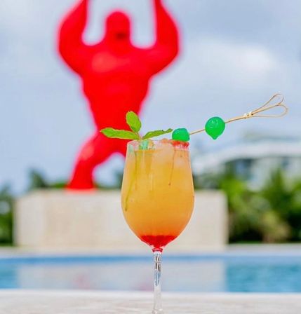 Tropical poolside cocktail in a stemmed glass — orange-red layered drink with mint and two green cherries on a skewer, ice glistening, blurred pool and large red sculpture in the background