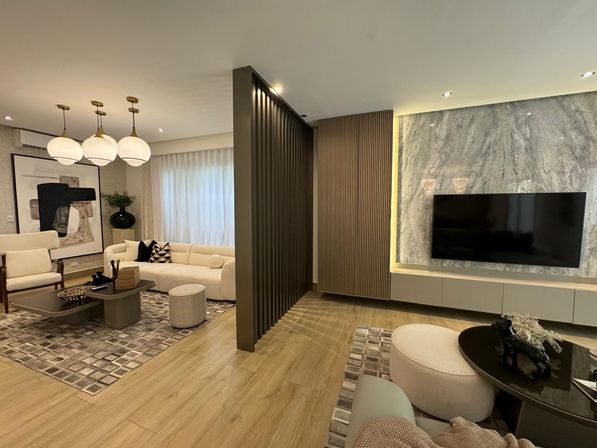 Sleek, cozy modern living room with neutral sofas, round pendant lights, wooden slat divider, marble TV feature wall, light wood floors and geometric area rugs.