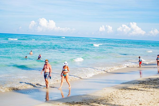 Tropical beach scene with turquoise ocean, blue sky and fluffy clouds, swimmers and beachgoers strolling along the wet sandy shoreline.