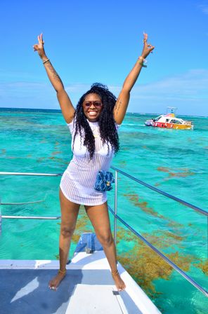 Smiling woman with curly hair and sunglasses on a boat deck, arms raised over bright turquoise tropical sea with a small colorful boat in the distance.