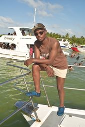 Man in sunglasses and baseball cap leaning on a boat railing at a crowded tropical boat party, palm-lined shore, multiple yachts and swimmers in green water under a sunny blue sky.