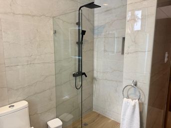 Sleek marble-look tiled bathroom with frameless glass walk-in shower, matte black rain and handheld shower fixtures, wood-look floor, toilet and towel ring with white towel.