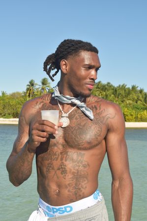 Tattooed shirtless man with braided hair and a bandana neck scarf holding a plastic cup, standing in shallow turquoise water with palm trees and a sandy tropical beach under a clear blue sky.