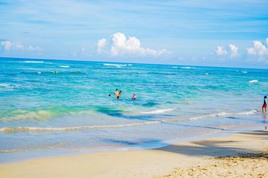 Sandy tropical beach with turquoise sea, gentle waves, people wading in shallow water and puffy white clouds