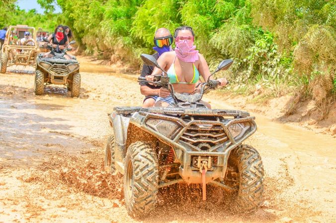 Couple wearing bandanas riding a muddy ATV through a tropical off-road trail, splashing mud as other ATVs follow on an adventure tour.
