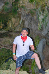 Man in white t-shirt, floral shorts, black cap and red bandana giving a thumbs-up while posing on rocks inside a moss-covered limestone cave, adventure travel scene.