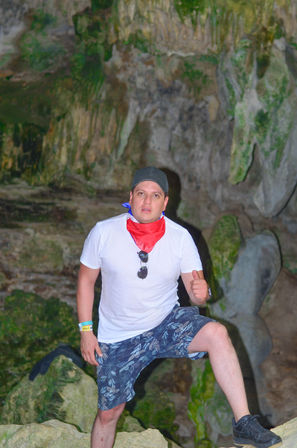 Man in white t-shirt, floral shorts, black cap and red bandana giving a thumbs-up while posing on rocks inside a moss-covered limestone cave, adventure travel scene.