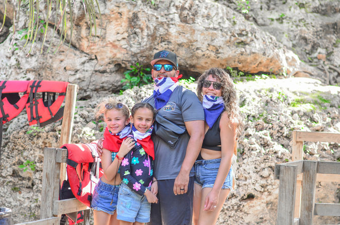 Smiling family of four posing on a tropical rocky shoreline, wearing colorful neck bandanas and sunglasses with life jackets on a wooden rail nearby