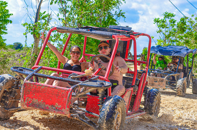 Smiling passengers in a red off-road dune buggy splashing mud on a sunny tropical dirt trail, with another buggy following behind