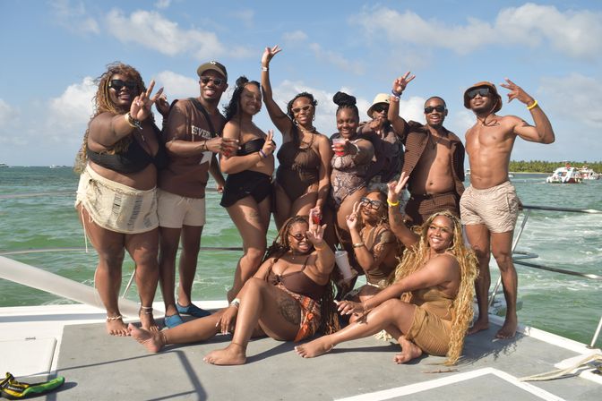 Cheerful group of people in swimwear posing with peace signs and drinks on the deck of a boat over turquoise tropical waters near a palm-lined coastline.