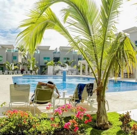 Tropical resort-style pool with central fountain, palm tree and bright bougainvillea, sun loungers with a person relaxing in front of modern apartment-style buildings.
