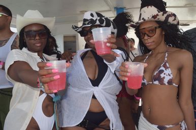 Three people in cowboy hats and bikinis holding plastic cups of pink cocktails, posing and cheering on a daytime boat party.