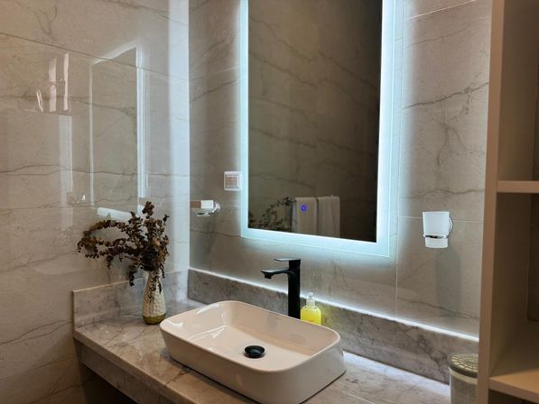 Sleek marble bathroom vanity with glowing LED mirror, white rectangular vessel sink, matte black faucet, yellow soap dispenser and a vase of dried branches